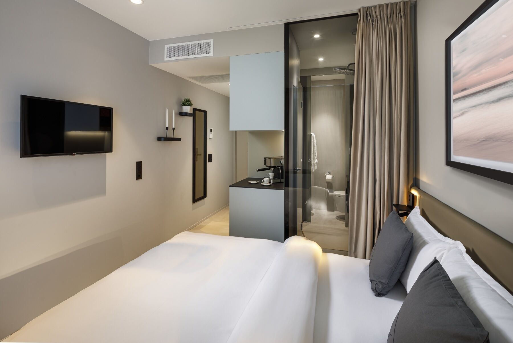 A modern hotel room with a large bed, a flat-screen TV, a small kitchenette, and a bathroom visible through a glass door.
