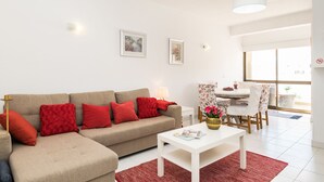 Ein modernes Wohnzimmer mit einem beigefarbenen Ecksofa, roten Kissen, einem weißen Couchtisch und einem Essbereich mit Tisch und Stühlen.