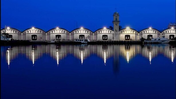 A waterfront building with multiple arched structures and a central tower, reflecting on calm water. - Biarritz Apartments (Gandia)