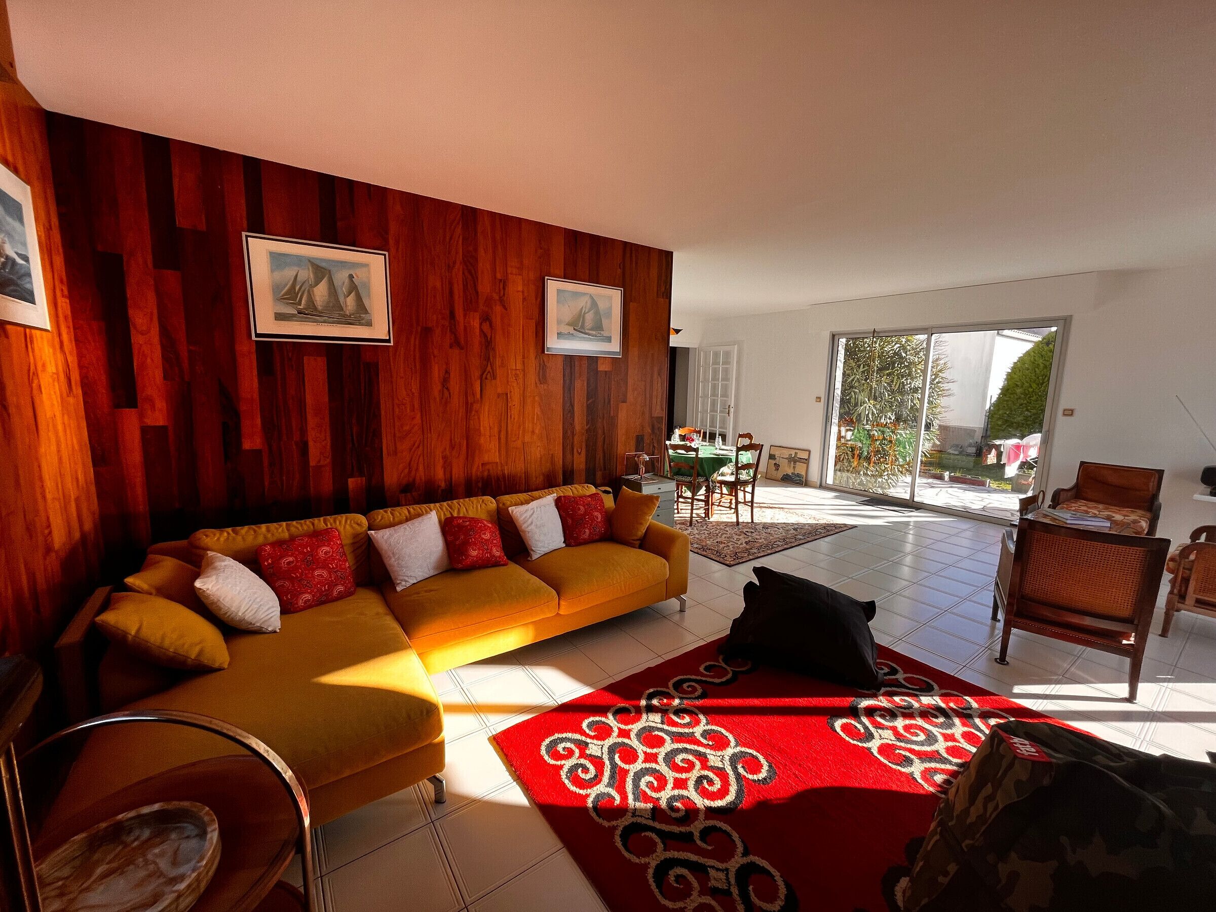 A living room with a wooden wall, a yellow sofa, a red patterned rug, and a dining area with a table and chairs.