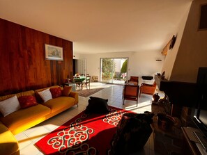Een ruime woonkamer met een gele bank, een rood vloerkleed met patroon, en een eethoek met een tafel en stoelen.