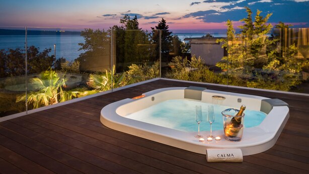A hot tub with a glass and champagne bottle, surrounded by wooden decking and a glass barrier, overlooking a body of water and trees at sunset. - Villa Calma I (Starigrad)