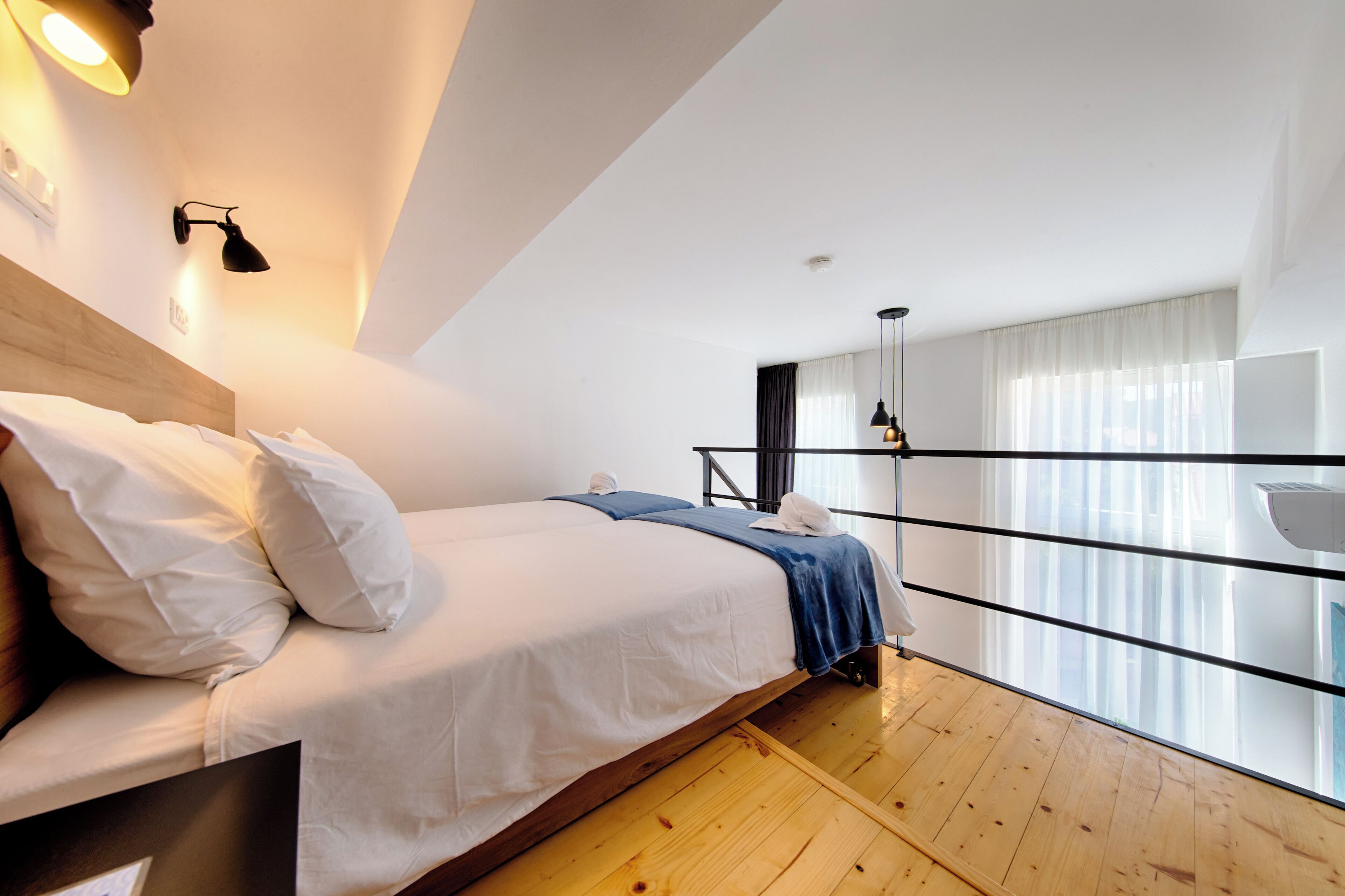 A modern hotel room with a wooden floor, a bed with white bedding, a blue towel, and a large window with curtains.