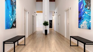 A hallway with wooden flooring, white walls, and two black benches. There are two large abstract paintings on the walls and a potted plant in the center. - 1901-on-sea (Vis)