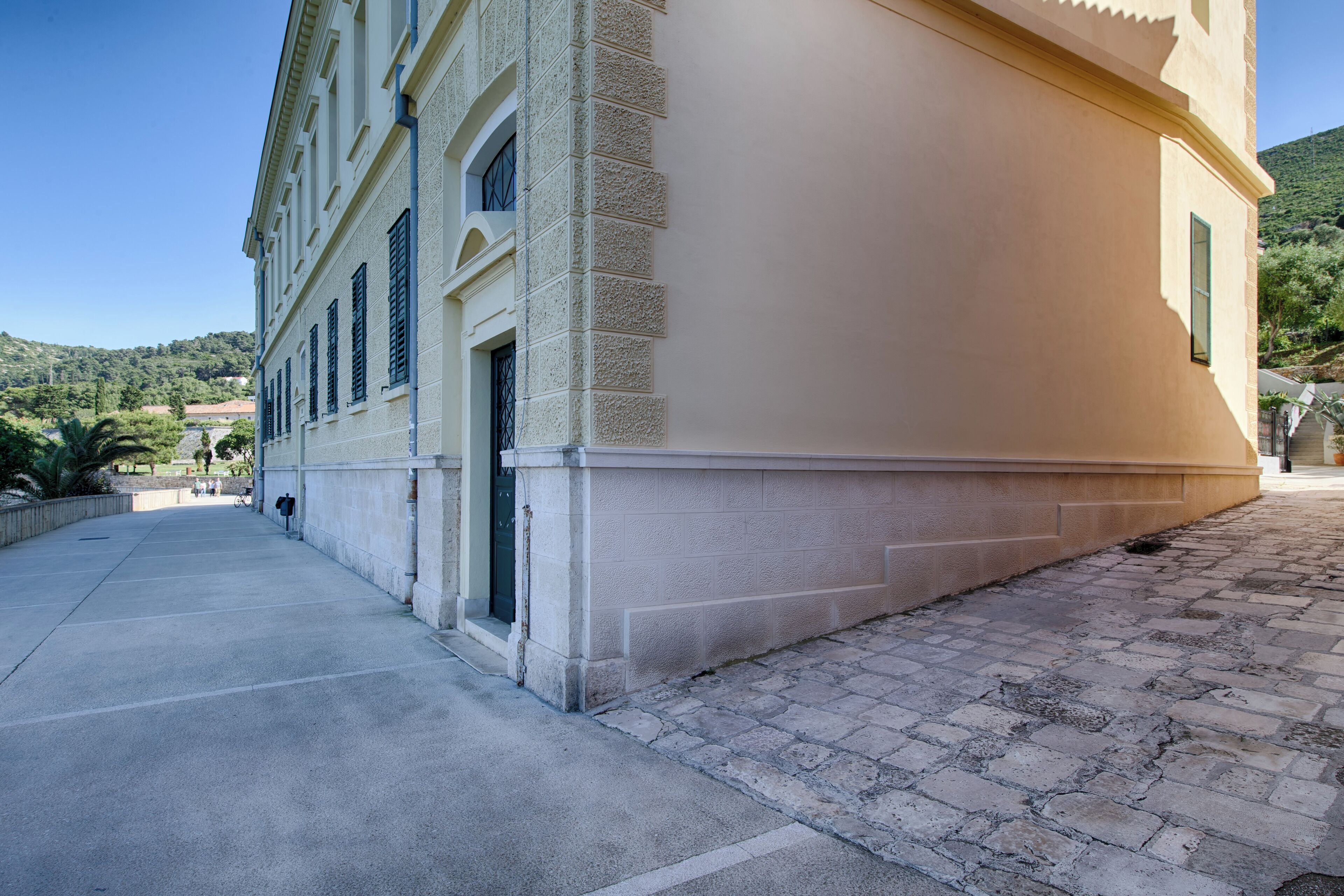 A building with a large, beige facade, a paved walkway, and a view of a hilly landscape in the background.