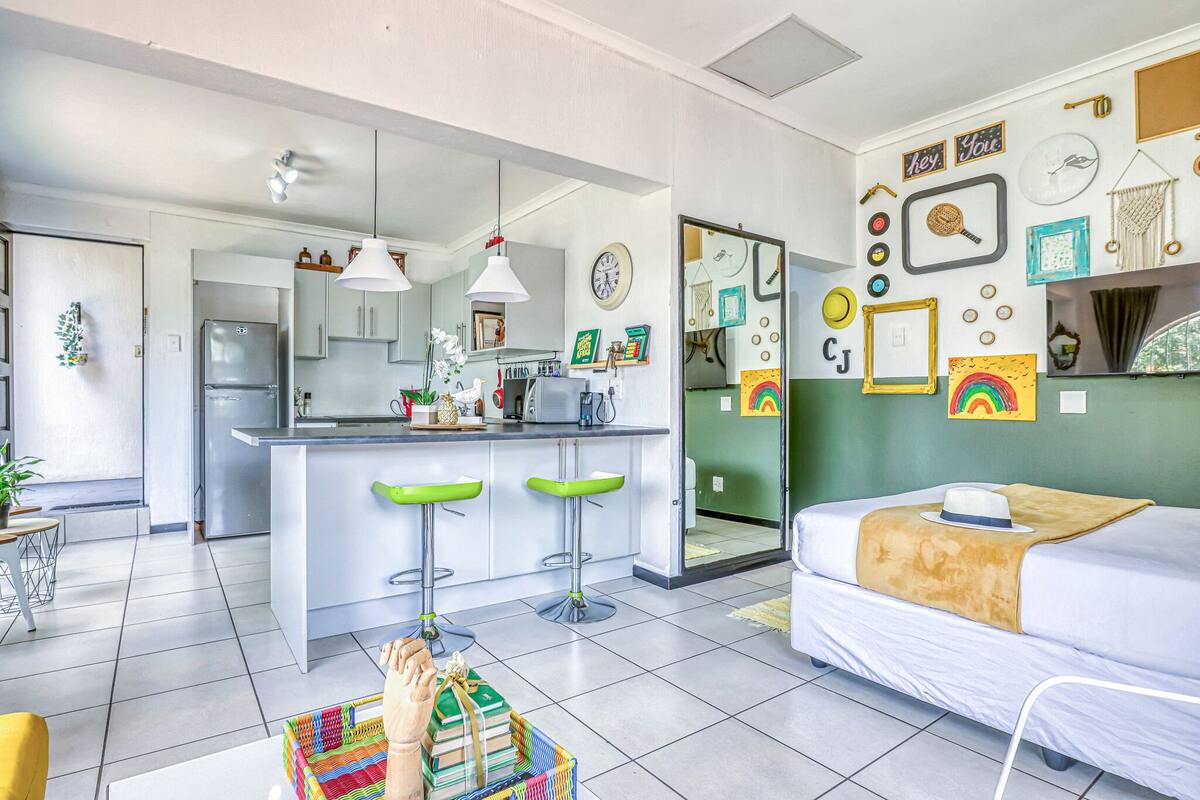 Una cocina moderna con una isla central, taburetes de bar y un comedor. Hay una cama con una manta blanca y marrón, y una pared decorada con varias fotos enmarcadas y un reloj.