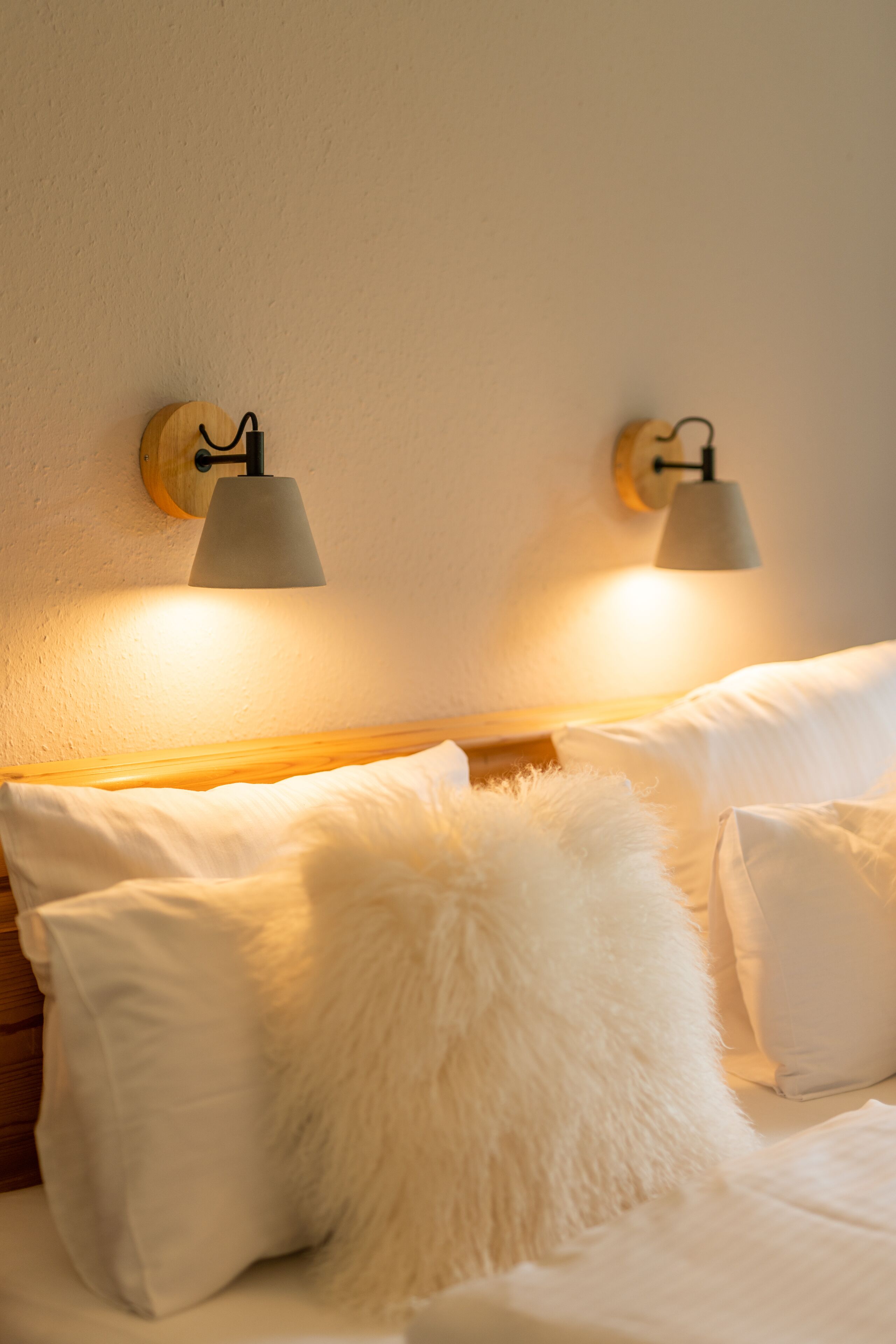 Cama com roupa de cama branca e almofadas fofas, duas luminárias fixadas na parede e cabeceira de madeira.
