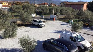 A parking area with several cars, a white van, and a yellow vehicle. There are buildings in the background with a mountainous landscape. - BRAND NEW APARTMENT JUST MINUTES FROM THE SEA (Sarzana)