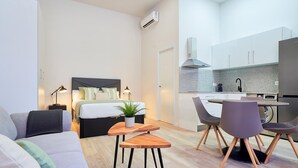 Un apartamento moderno con dormitorio, sala de estar y cocina. El dormitorio tiene una cama con cabecera, lámparas de noche y una planta en maceta. La sala de estar incluye un sofá, una pequeña mesa de madera y una silla. La cocina cuenta con alacenas blancas, una estufa y un fregadero.