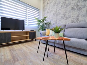 A modern living room with a flat-screen TV, wooden entertainment unit, grey sofa, and a small table with a potted plant.