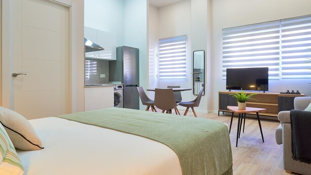Una habitación de hotel moderna con una cama, un escritorio pequeño con una silla, un televisor y una zona de cocina con lavadora.