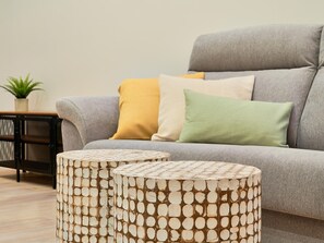 Un sofá gris con tres cojines en amarillo, blanco y verde, y dos mesitas redondas con un diseño tejido.