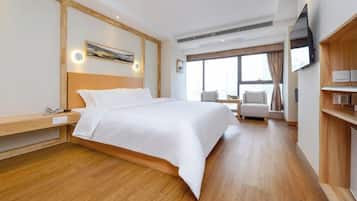 A hotel room with a large bed, wooden flooring, a desk with a telephone, a TV on the wall, and a window with curtains.