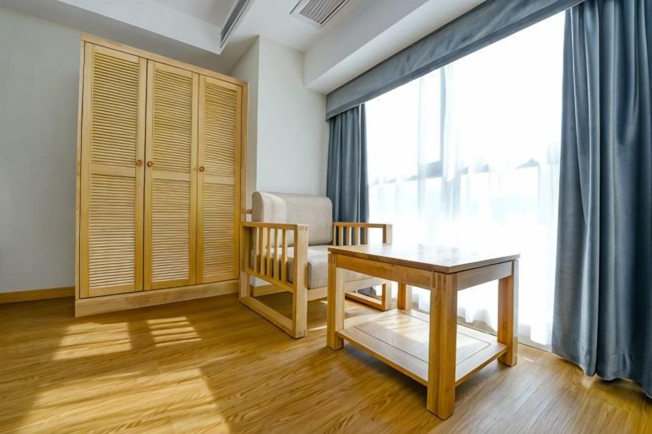 A room with a wooden wardrobe, a small table, a chair, and a large window with curtains.