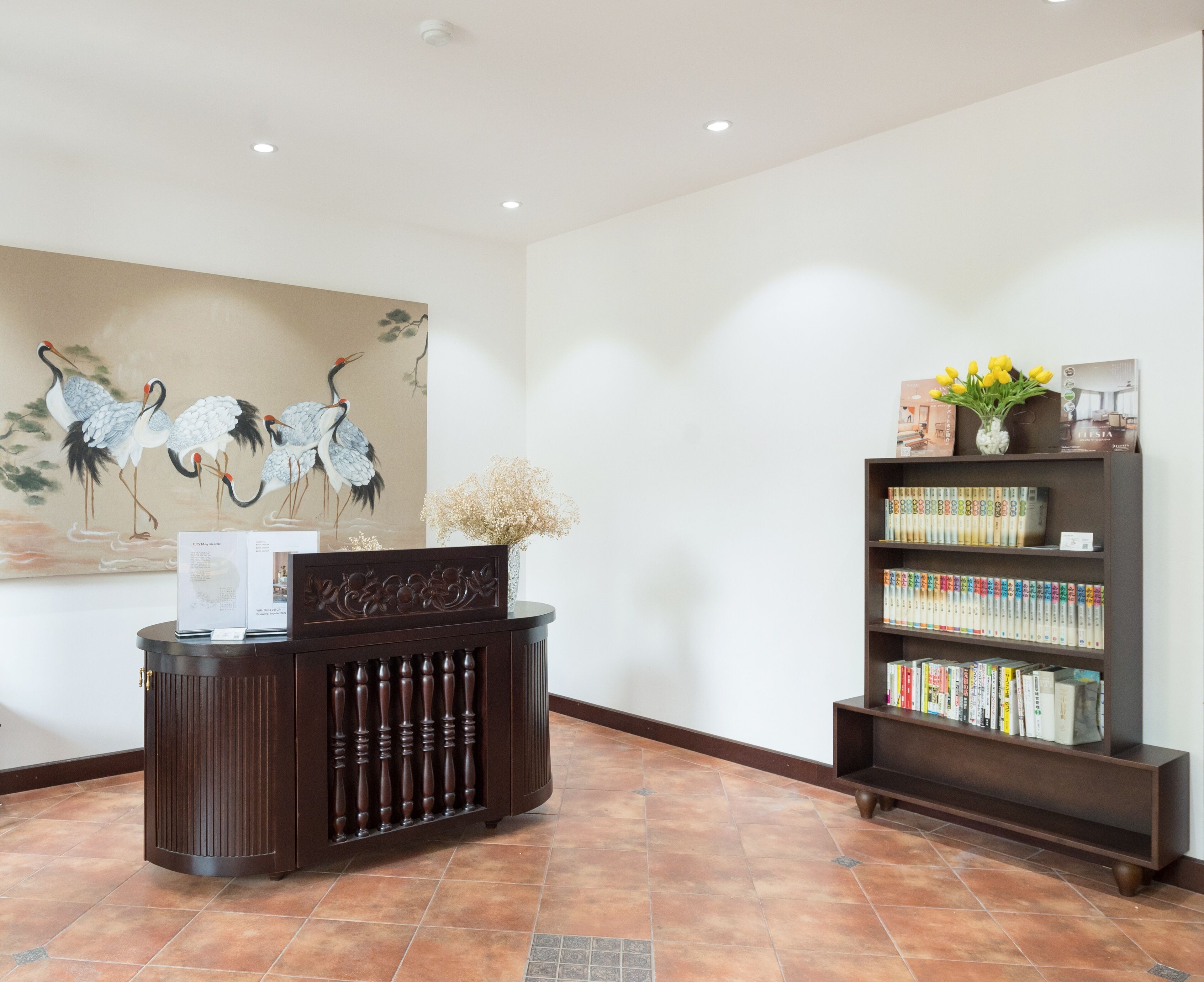 A reception area with a dark wooden desk, a bookshelf filled with books, a vase with yellow flowers, and a painting of cranes on the wall.
