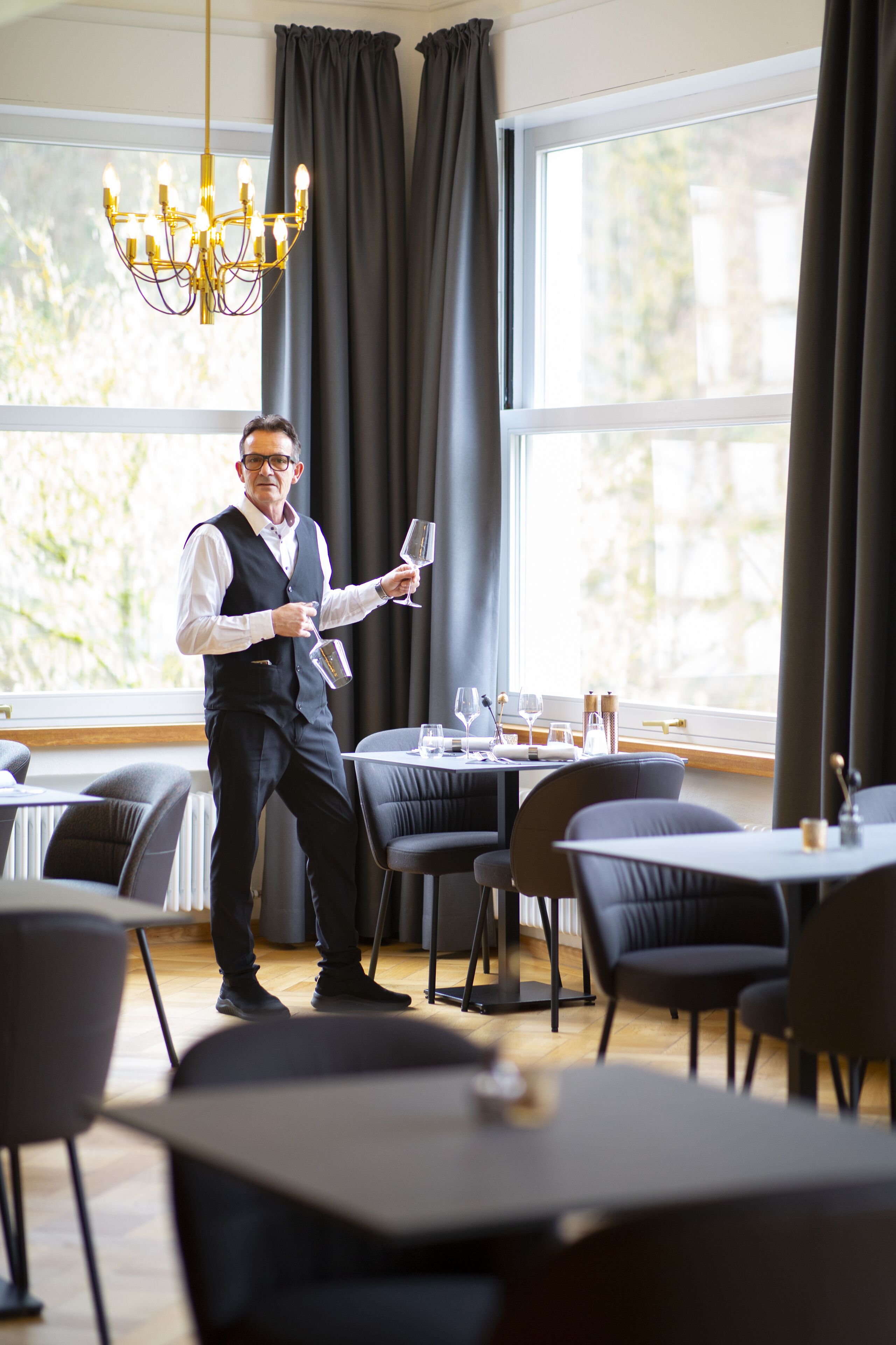 A waiter holding a wine glass in a well-lit dining area with large windows, dark curtains, and modern furniture.