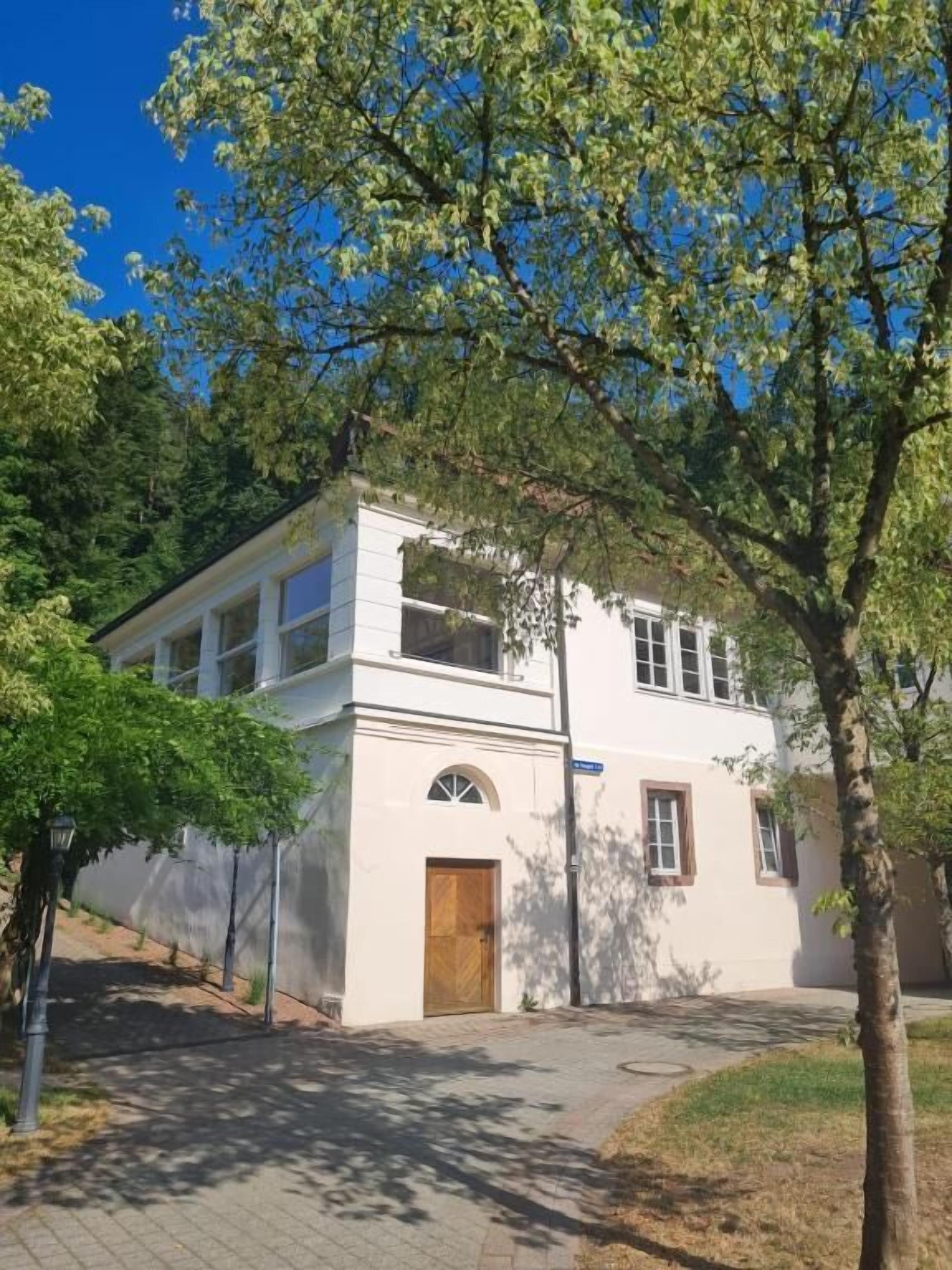 Ein zweigeschossiges Gebäude mit weißer Fassade, einer Holztür und einem kleinen Rundbogenfenster.