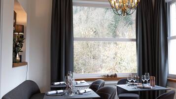 Un coin repas avec une table dressée pour quatre personnes, un lustre et une vue sur les arbres à travers de grandes fenêtres.
