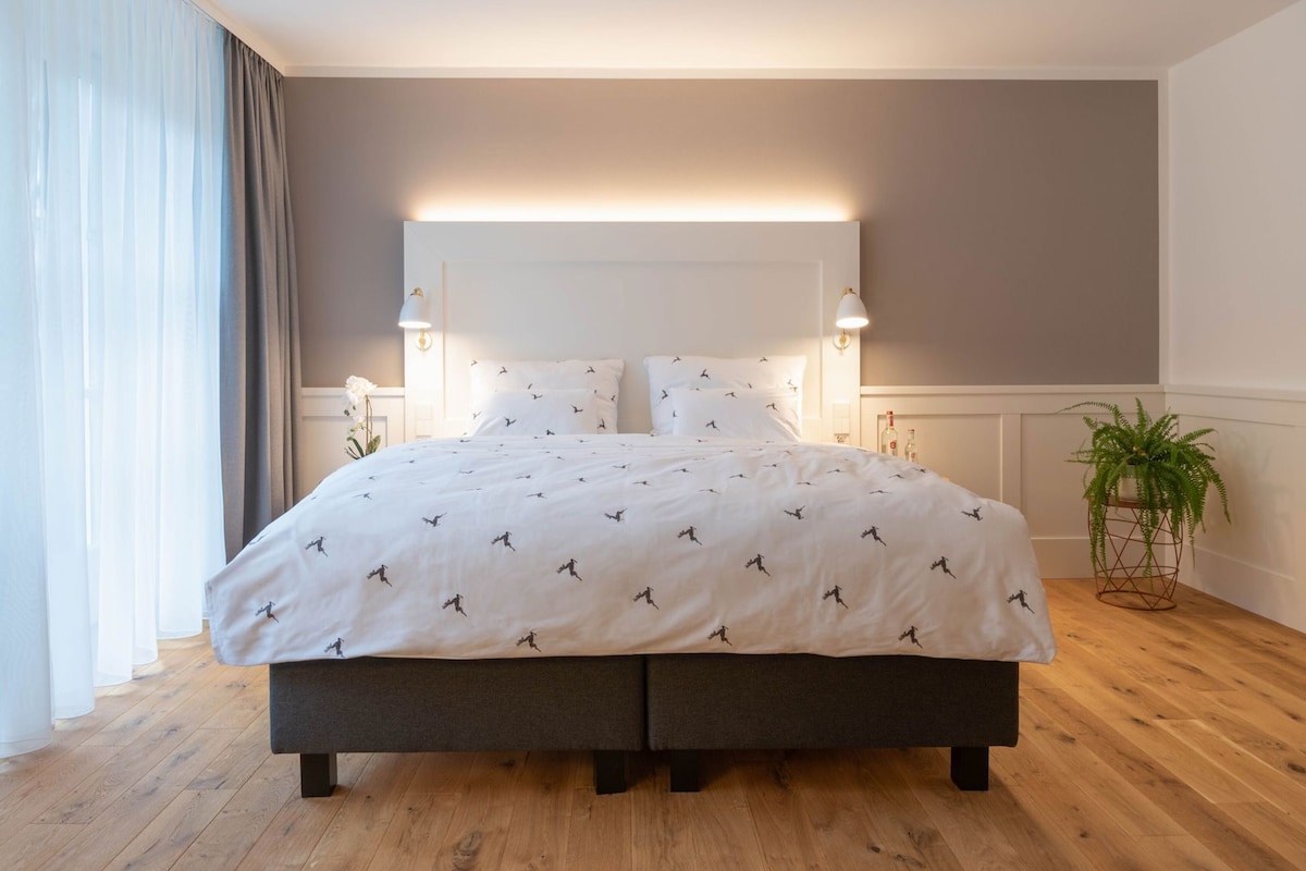 A modern bedroom with a large bed, two bedside lamps, a potted plant, and a view of the outdoors.