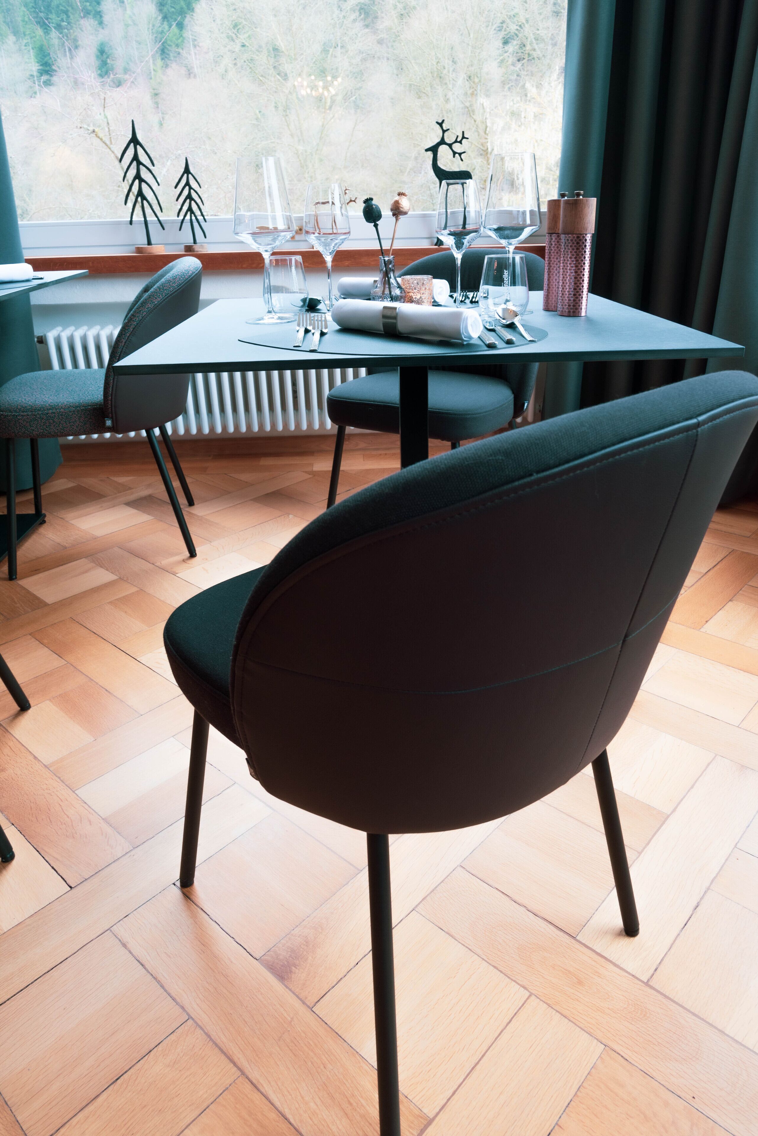 Comedor con una mesa lista para dos personas, sillas y vista a árboles por la ventana.