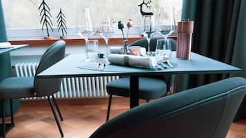 Comedor con una mesa lista para dos personas, sillas y vista a árboles por la ventana.