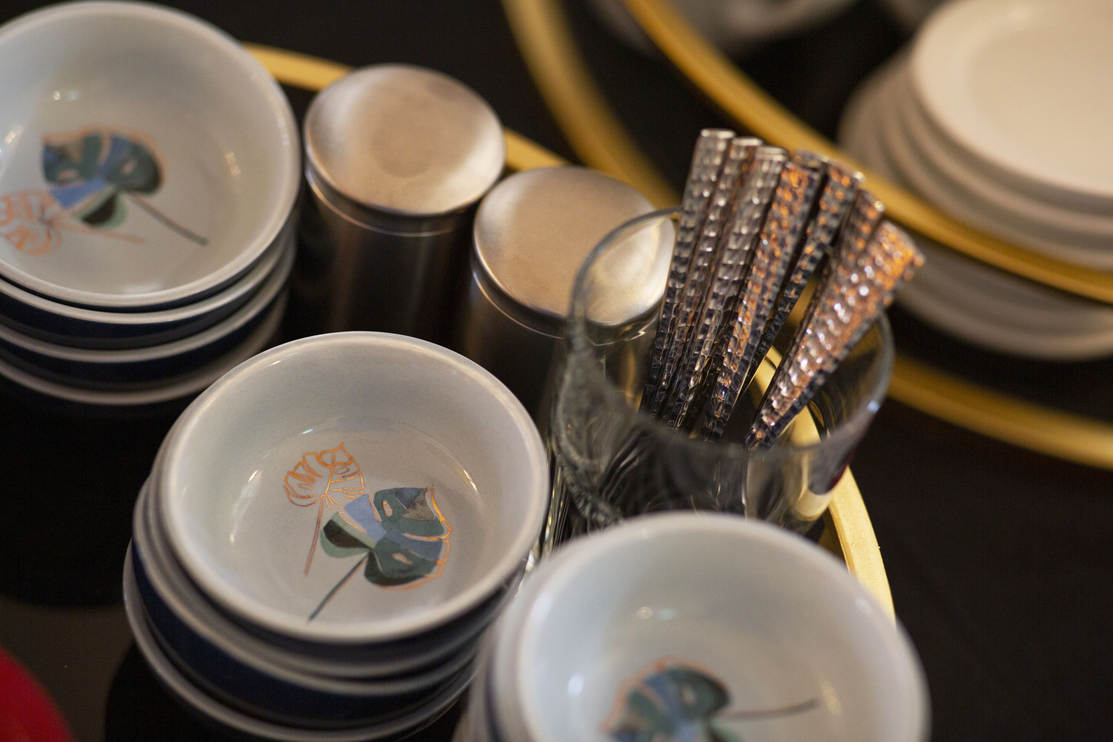 A stack of white plates with a leaf design, a clear glass container with silverware, and a gold-colored tray.