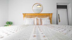 Una cama bien hecha con cabecera de madera, un espejo redondo encima y una pequeña planta en maceta a la izquierda.