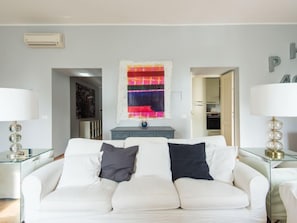 Una sala de estar moderna con un sofá blanco, dos mesas auxiliares con lámparas, un cuadro abstracto de colores y una cocina visible a través de una puerta abierta.
