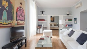 A modern living room with a flat-screen TV, white sofa, and a glass coffee table. There is a bookshelf with books and a painting on the wall. - La Casa dell'Arte by Interhome (Rome: Historical City Center)