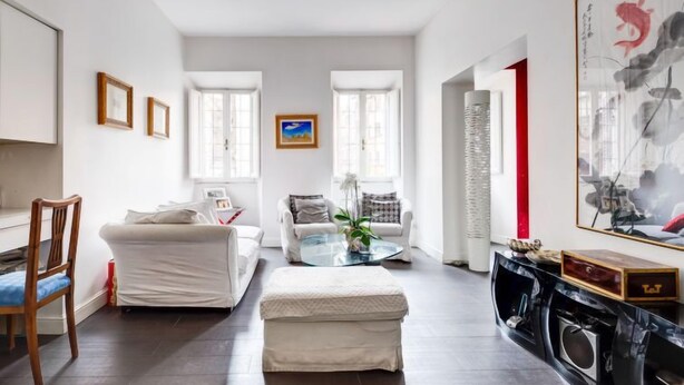 Una sala de estar moderna con un sofá blanco, una mesa de centro de vidrio y un pequeño otomaní. Hay cuadros enmarcados en la pared, una pintura y una obra de arte abstracta de gran tamaño.