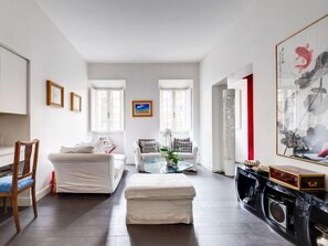 A modern living room with a white sofa, a glass coffee table, and a small ottoman. There are framed pictures on the wall, a painting, and a large abstract artwork.