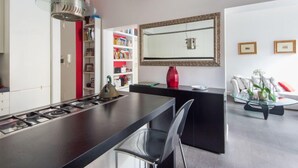 Una cocina moderna con estufa a gas, un comedor con mesa negra y una silla de vidrio. Hay una estantería con libros de colores y un florero rojo sobre la encimera. Un espejo grande cuelga de la pared, y un sofá con cojines blancos se ve al fondo.