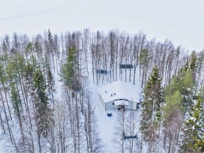 Vue aérienne d’une cabane entourée d’une forêt dense, le sol étant recouvert de neige.
