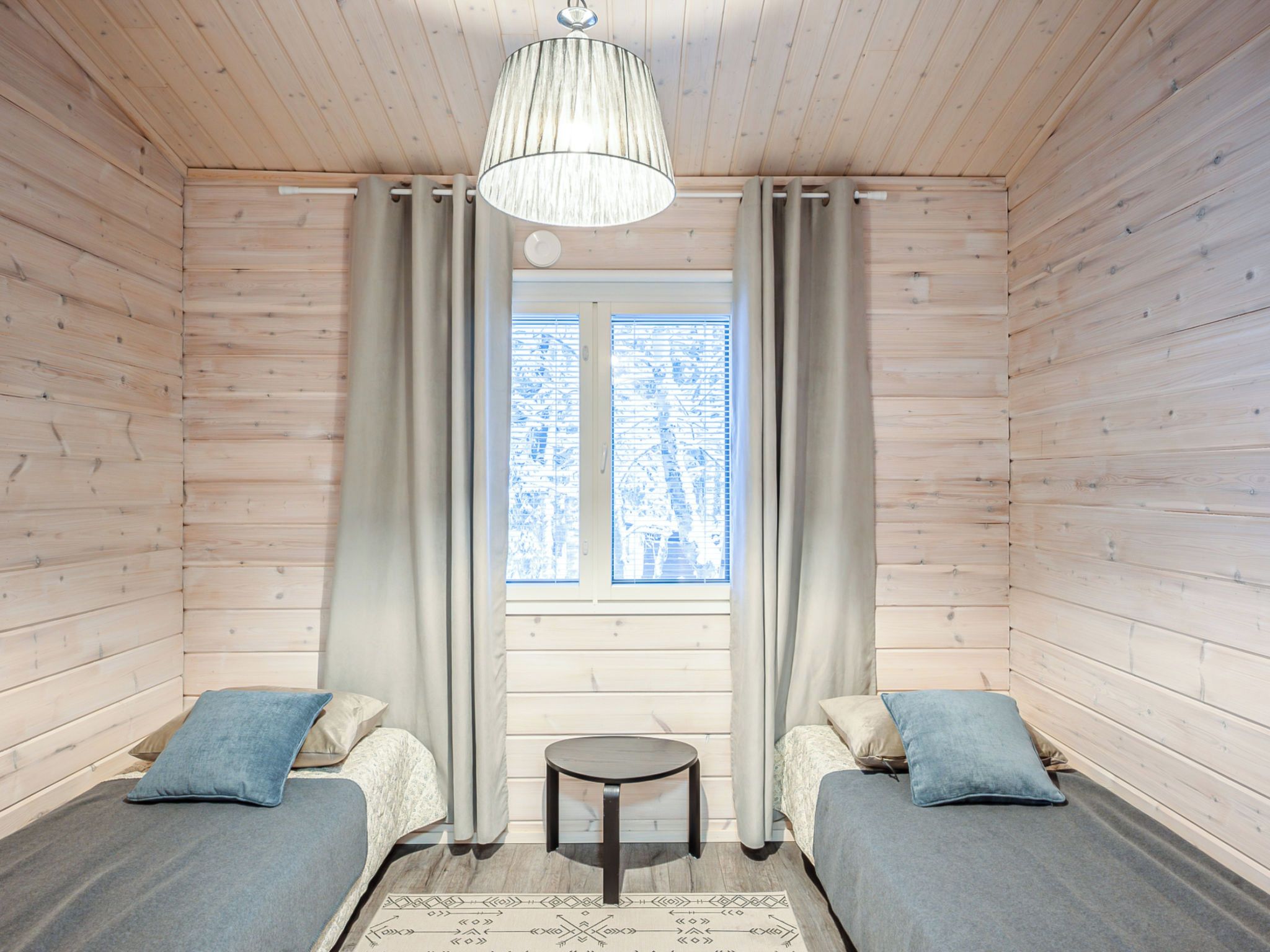 A room with wooden walls and a window with curtains. There are two beds with pillows and a small table in the center.
