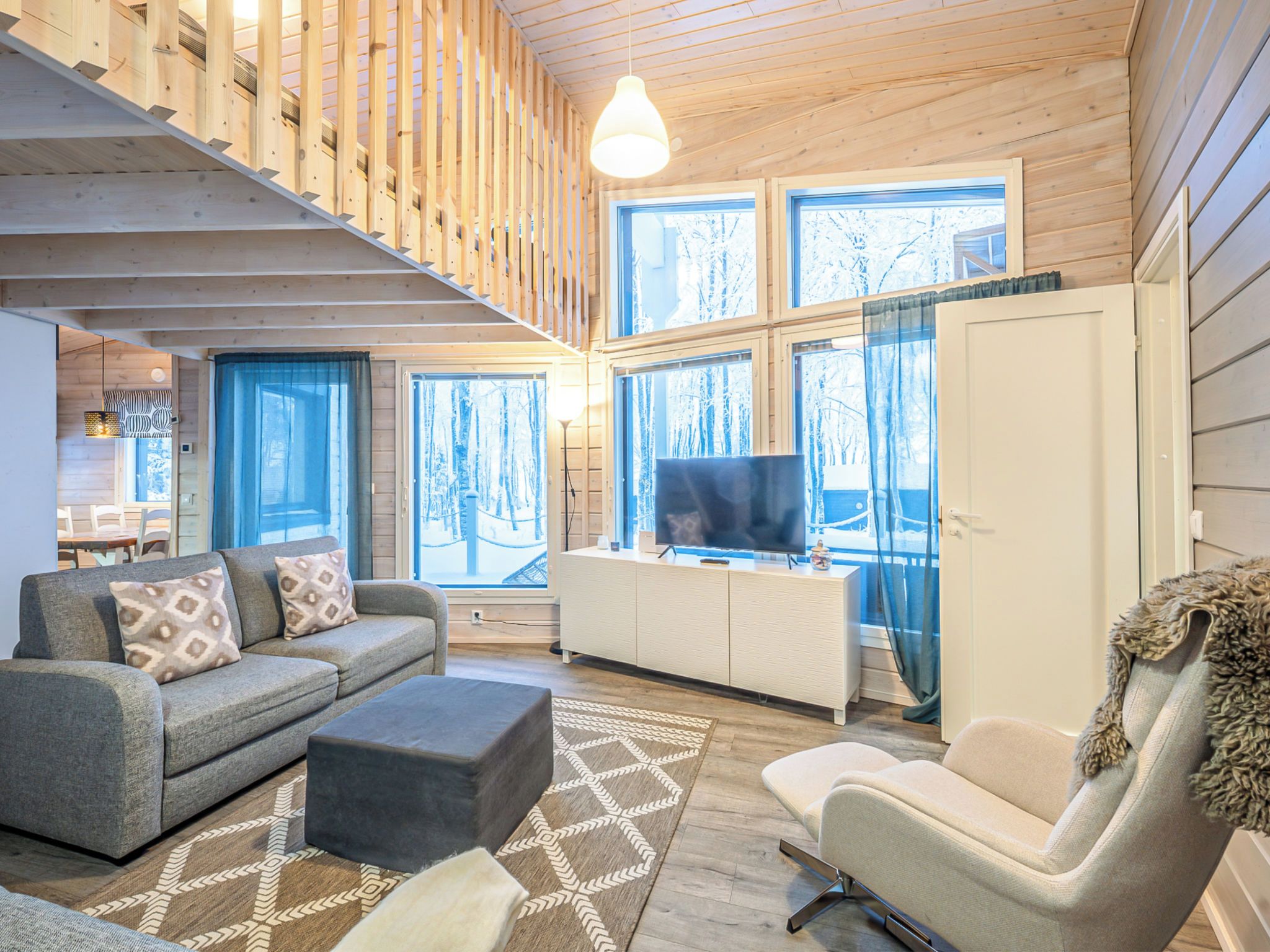A modern interior with a wooden staircase, a sofa set, a TV on a cabinet, and large windows with a view of trees.