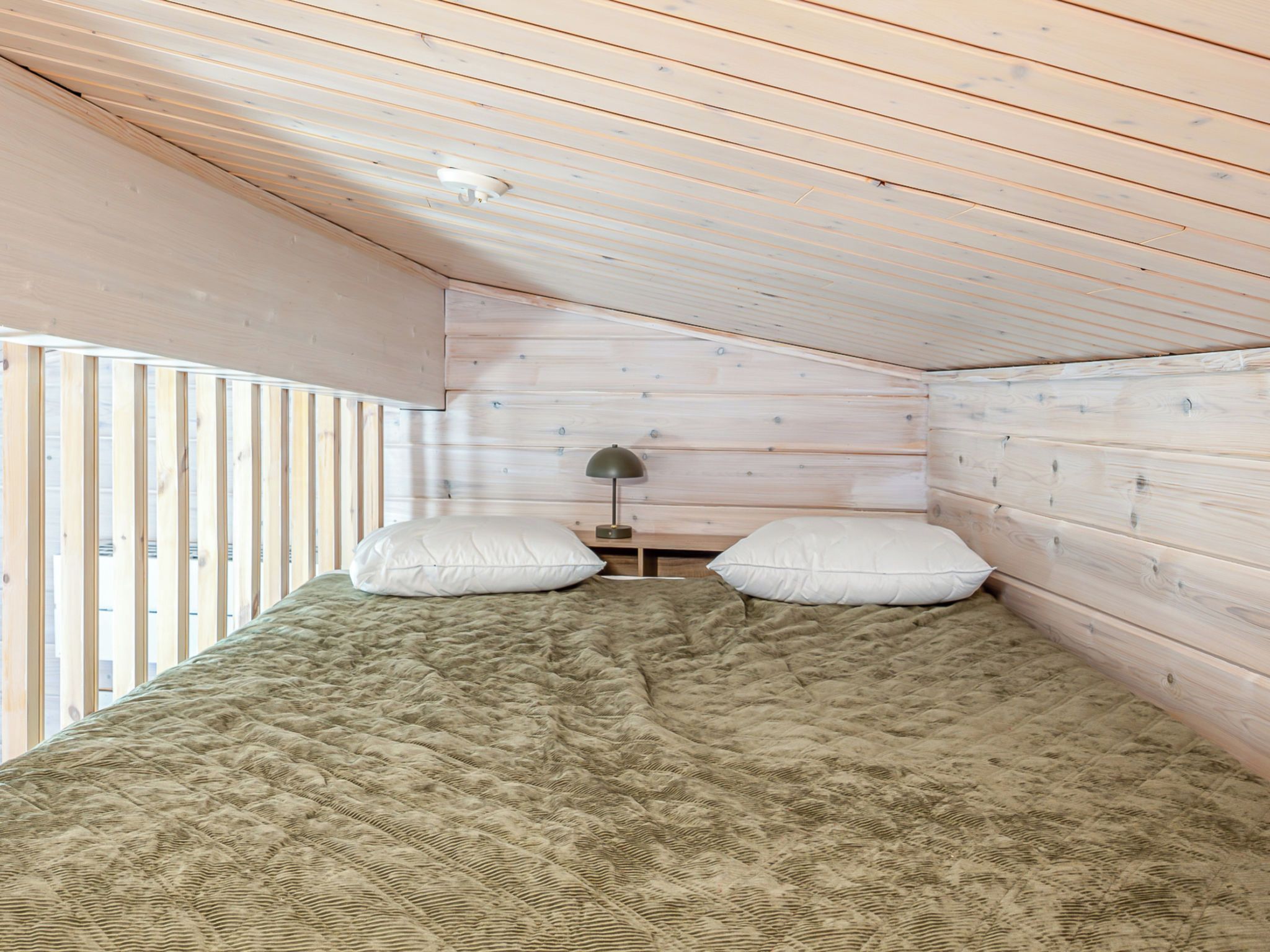 A small, cozy room with a sloped ceiling and wooden walls. There is a bed with a quilted blanket and two pillows. A small table with a lamp is against the wall.
