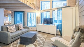 A modern interior with a wooden staircase, a sofa set, a TV on a cabinet, and large windows with a view of trees.