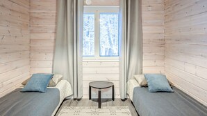 A room with wooden walls and a window with curtains. There are two beds with pillows and a small table in the center.
