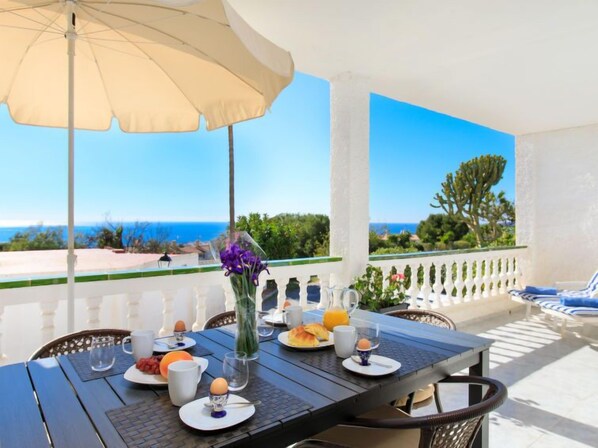 Una mesa de desayuno en un balcón con vistas al mar, barandilla blanca y un paraguas beige.
