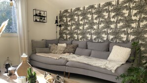 Een woonkamer met een grijze bank, een bijzettafeltje met decoratieve objecten, een muur met een boommotief en een raam met gordijnen.