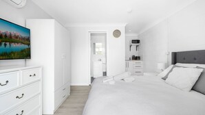 A modern bedroom with a large bed, a dresser, a television, and a kitchen area. - Beach Views on Excellent Studio 27 - Belle Escapes Jervis Bay (Vincentia)
