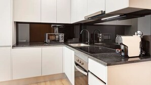 Una cocina moderna con gabinetes blancos, encimera negra, horno integrado, microondas, cafetera y pileta de acero inoxidable.