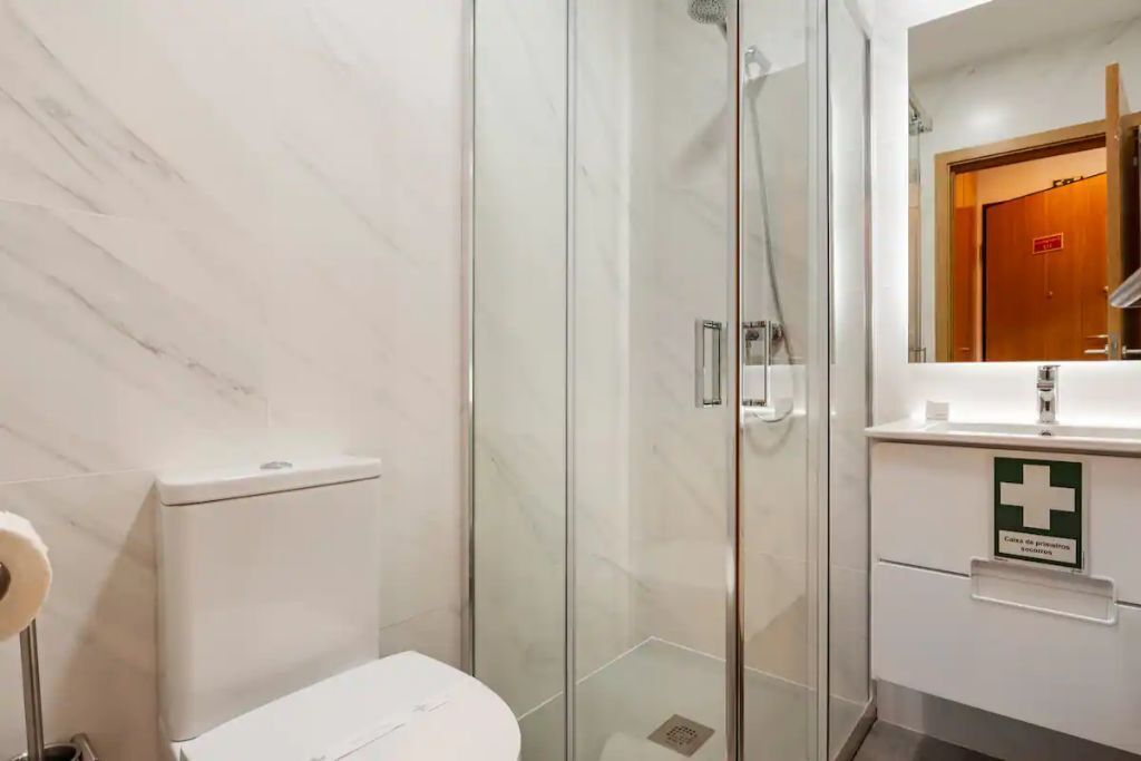 A bathroom with a glass-enclosed shower, a toilet, and a sink with a first aid sign.