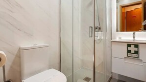Baño con ducha envuelta en vidrio, inodoro y lavamanos con un cartel de primeros auxilios.