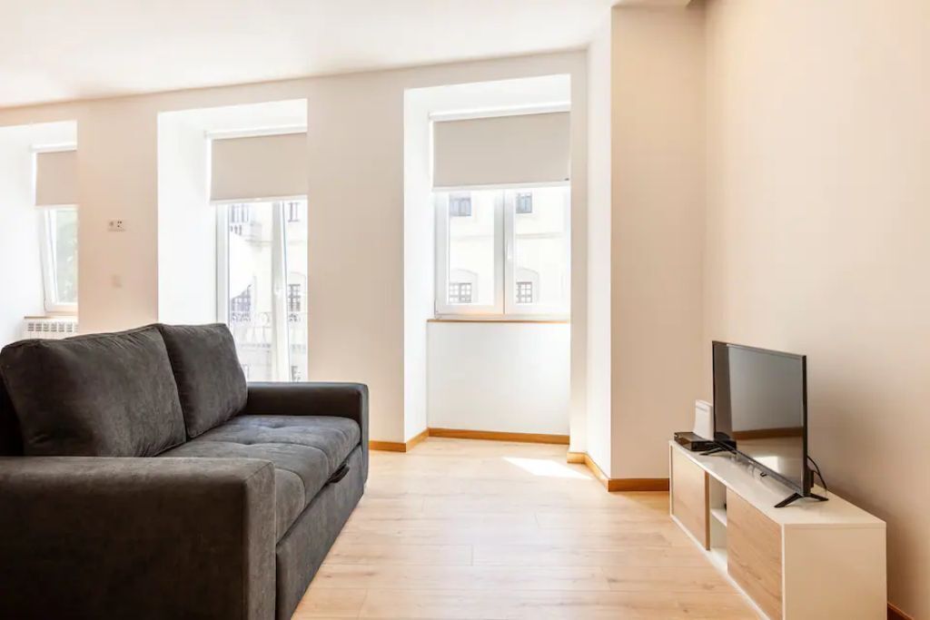Una sala de estar moderna con un sofá gris, un televisor de pantalla plana sobre un mueble de madera y grandes ventanas que ofrecen vistas a edificios.