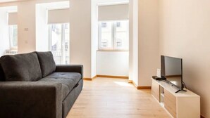 Una sala de estar moderna con un sofá gris, un televisor de pantalla plana sobre un mueble de madera y grandes ventanas que ofrecen vistas a edificios.