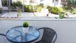 Uma mesa de vidro redonda com duas cadeiras, uma planta em vaso e dois copos de vinho numa varanda com vista para um jardim e edifícios.