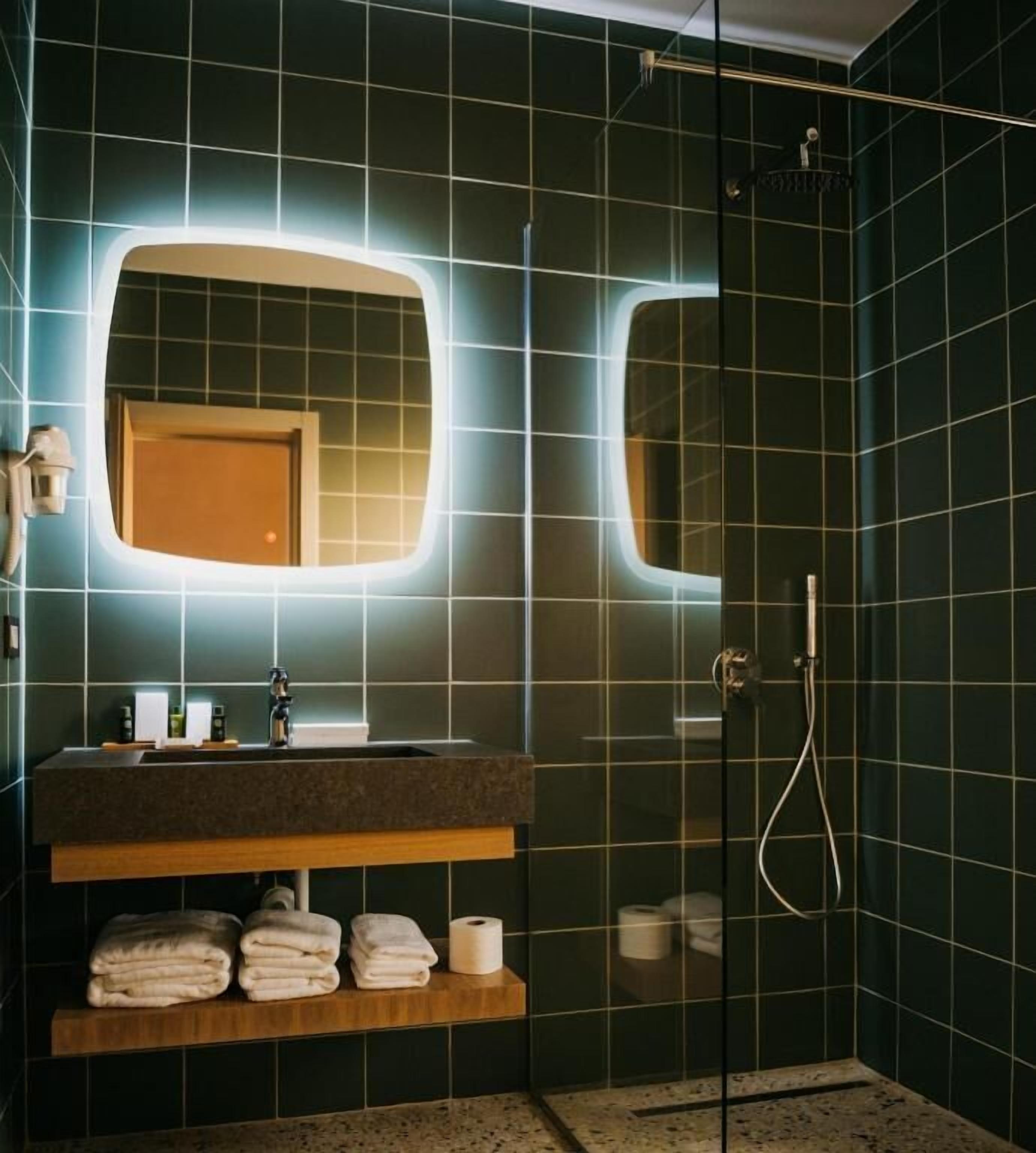 Un baño moderno con azulejos verde oscuro, un mueble de tocador de madera, un espejo grande y una ducha con mampara de vidrio.