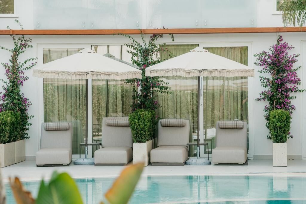 Area piscina con quattro lettini e due ombrelloni bianchi, circondata da piante in vaso e una struttura in vetro.