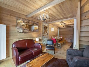 A cozy cabin interior with a wooden ceiling, a red leather sofa, a wooden coffee table, and a dining area with a table and chairs.